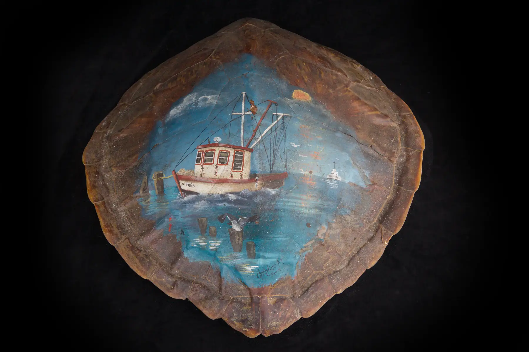 A turtle shell painted on the inside with a fishing boat on blue water, gulls overhead and mooring posts at the left