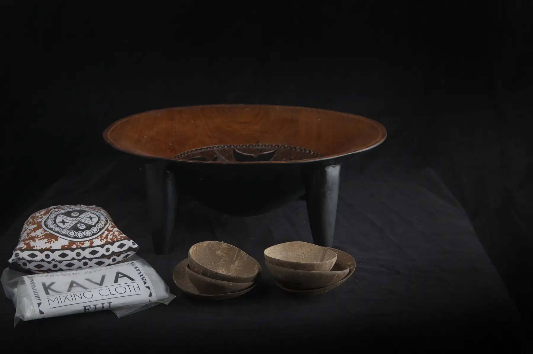 A carved wooden kava bowl on legs with several coconut-shell cups, a printed Fiji kava mixing cloth and a patterned cloth bundle