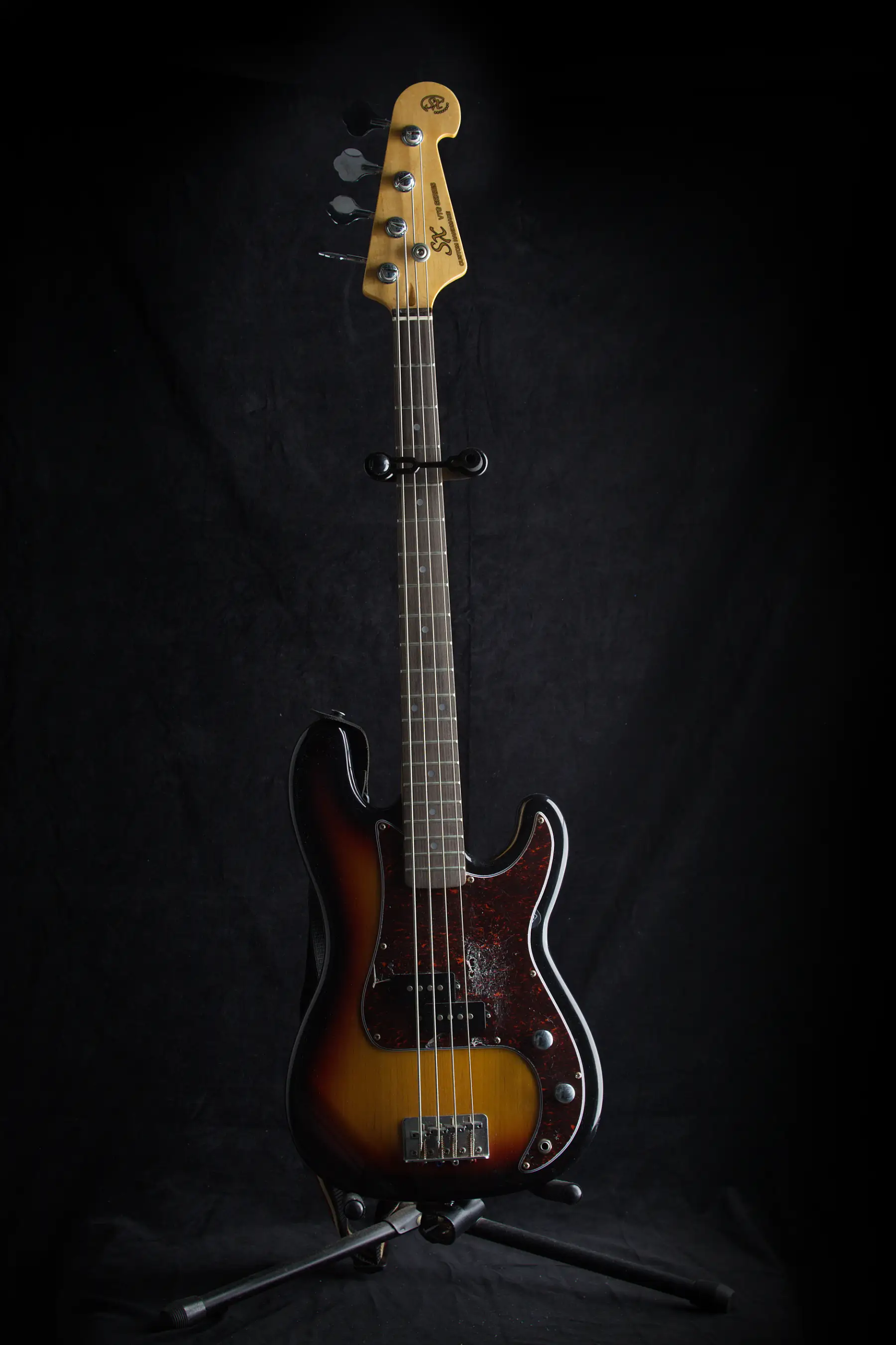 A four-string sunburst electric bass on a stand against black, with a maple neck, rosewood fingerboard and tortoiseshell pickguard